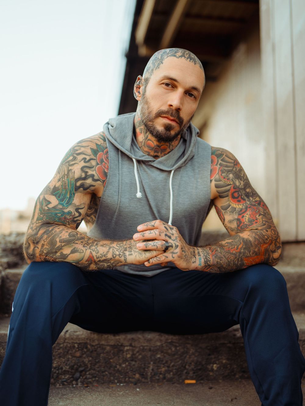 Tattooed person in gray hoodie and jeans sitting outdoors with colorful arm sleeve artwork visible.