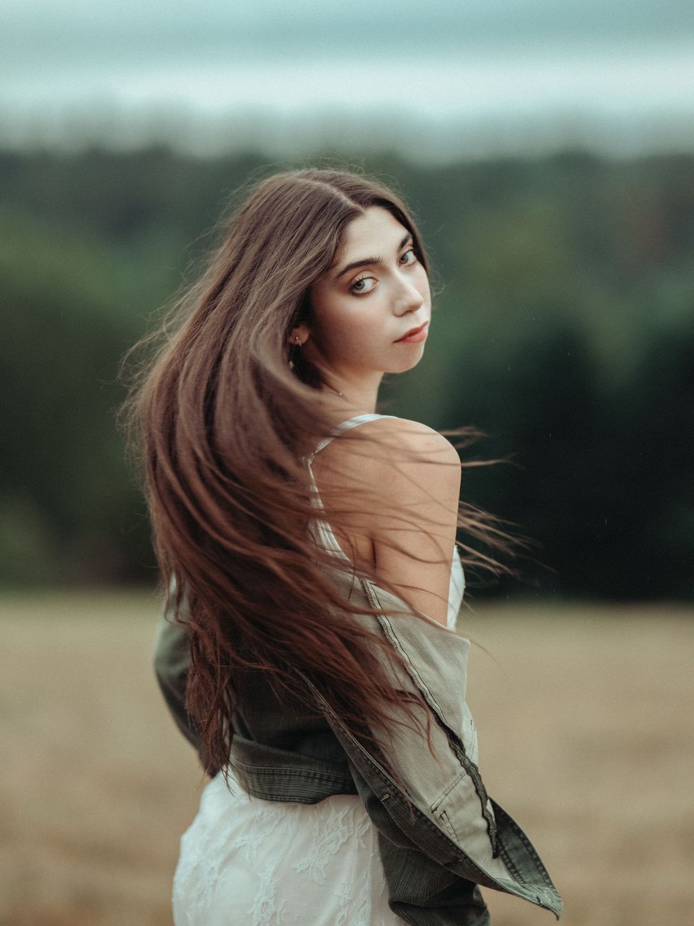 Woman with flowing brown hair twirls in a serene outdoor field on an overcast day wearing a light-colored top.