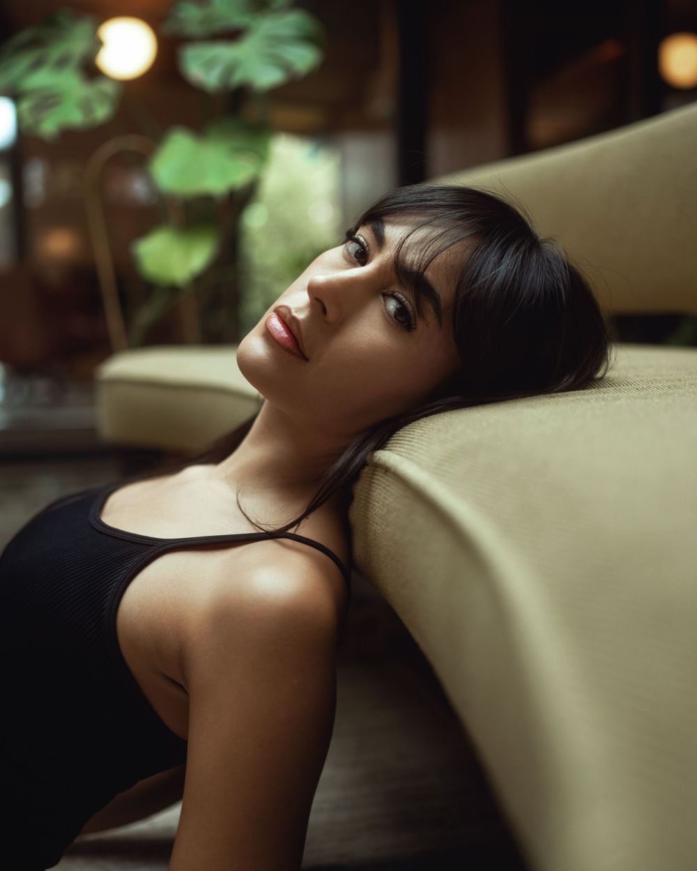 Creative editorial portrait in moody lighting with black top resting against beige couch, with green leaves and soft bokeh in background.