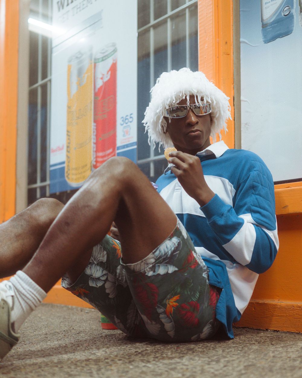 A person wearing a blue sweater and printed shorts sits casually against a wall with white hat and sunglasses.