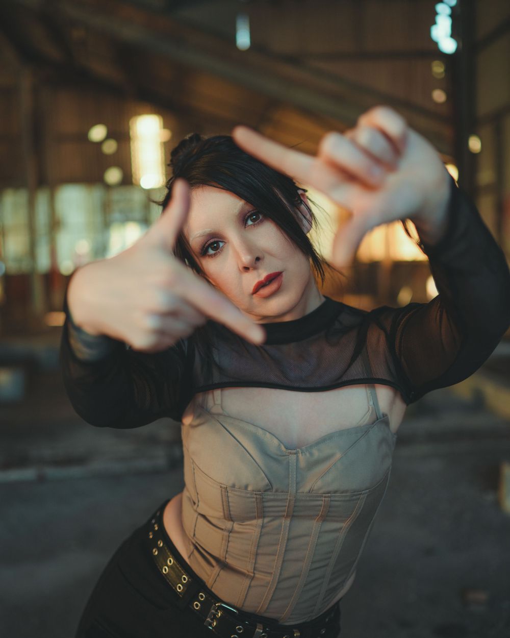 Alternative fashion model in sheer black top and corset poses artistically with hands framing face in moody lighting.