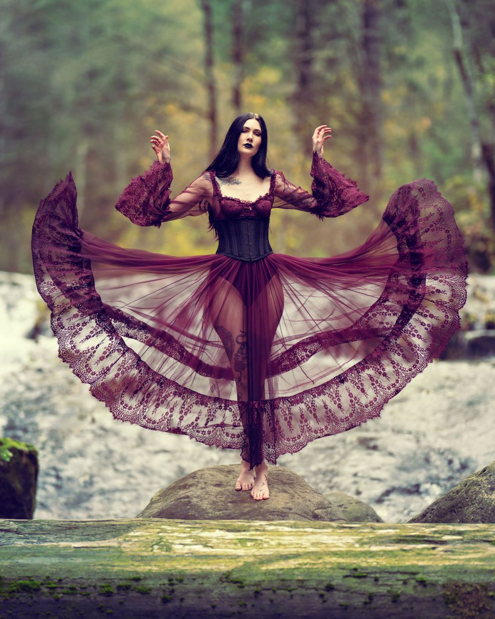 A whimsical figure in a flowing burgundy lace dress poses gracefully on a rock by a stream in a misty forest setting, artistic photoshoot.
