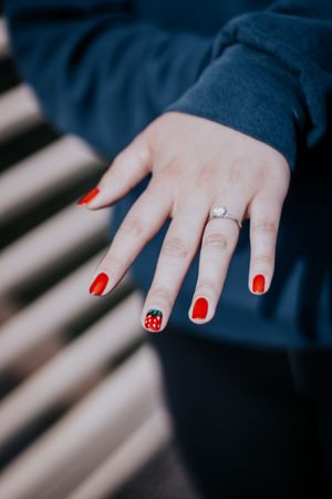 Close-up of a hand with bright red nail polish and a small engagement ring against a dark blue fabric background.