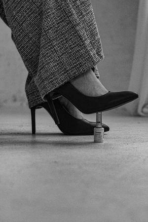 A black stiletto heel rests on a tiny bottle in an artistic black and white composition on a concrete floor.