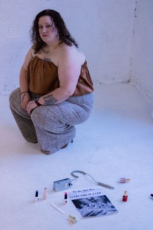 Figure in brown top and plaid pants poses in studio setting with various items scattered on white floor.