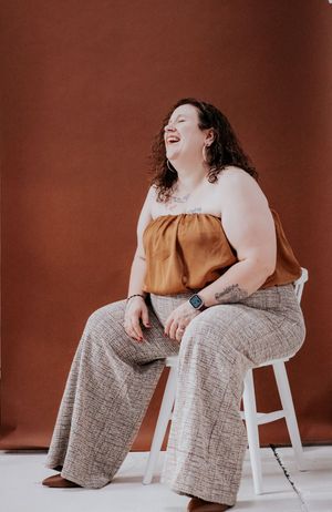 A joyful person in a brown tank top and textured wide-leg pants sits on a white stool against a terracotta backdrop.