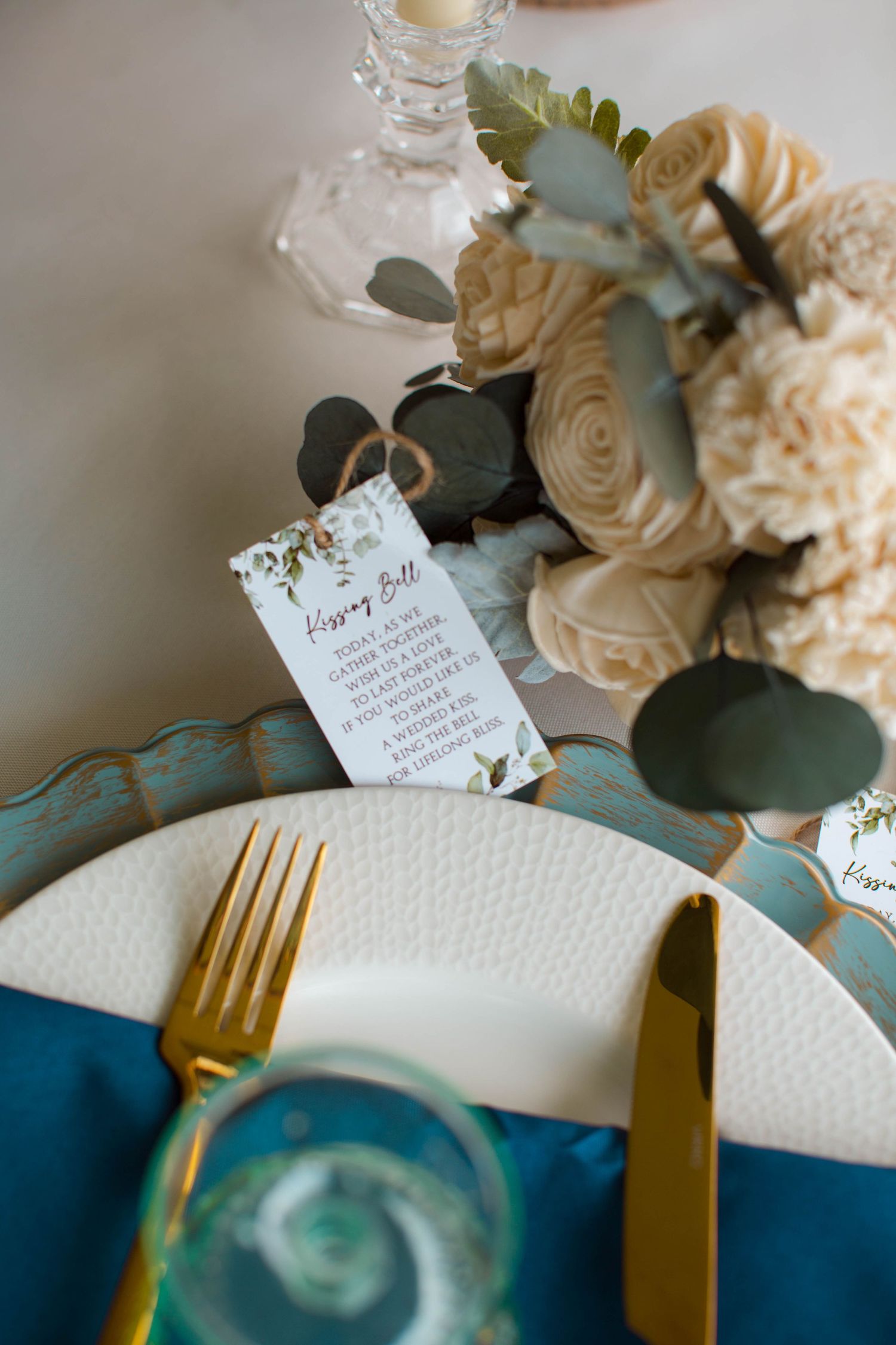 Elegant wedding place setting with cream roses, gold cutlery, and a menu card decorated with eucalyptus on teal linens.