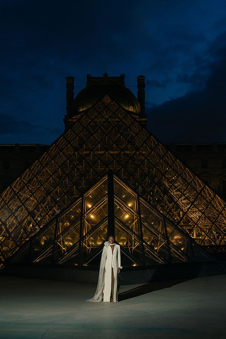 A person in white stands in front of the illuminated Louvre pyramid at night.