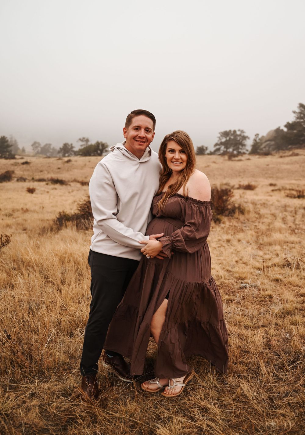 Couple poses together in field wearing brown dress and gray hoodie.