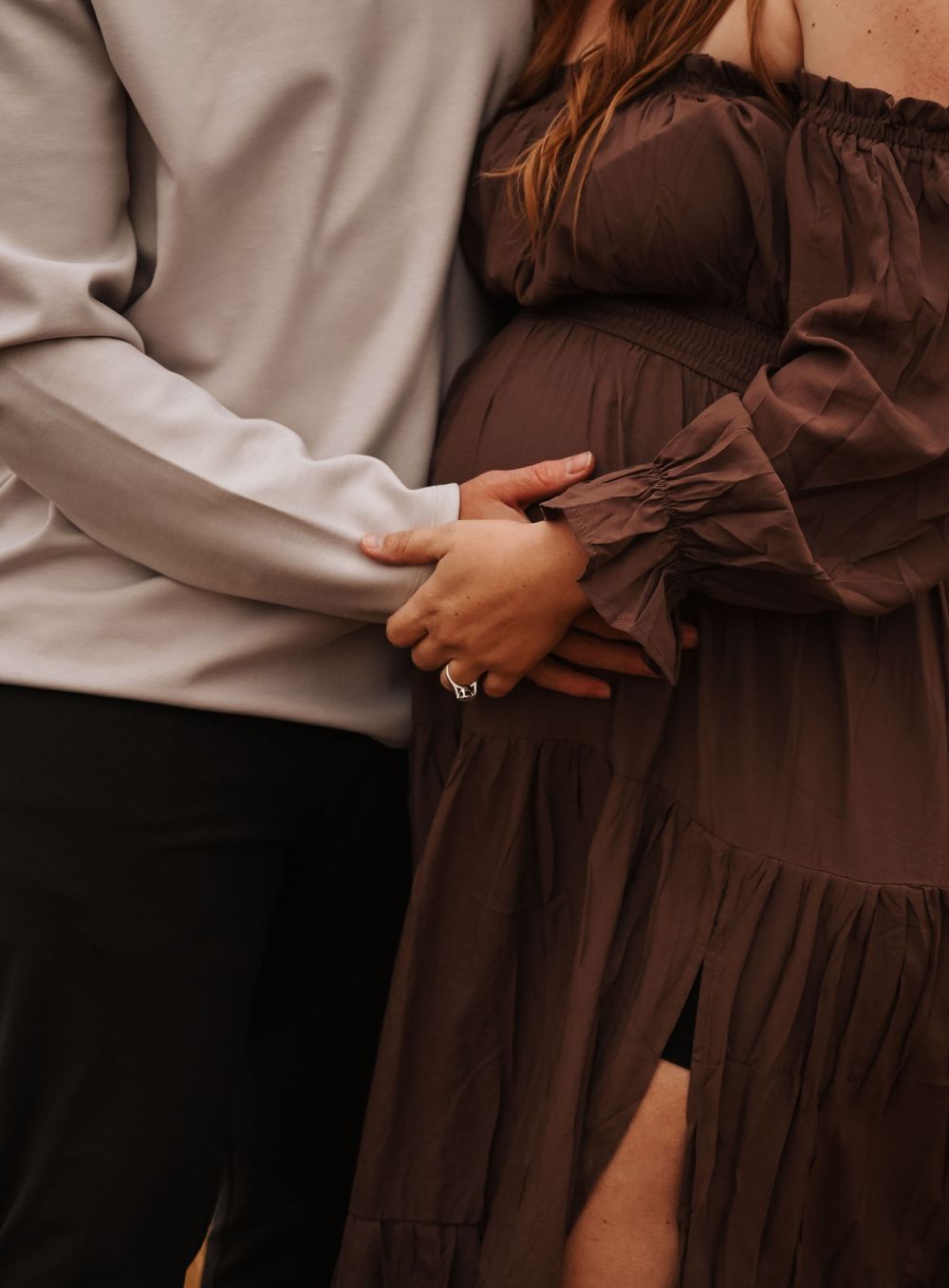 Close-up of couple embracing with hands on pregnant belly in brown dress.