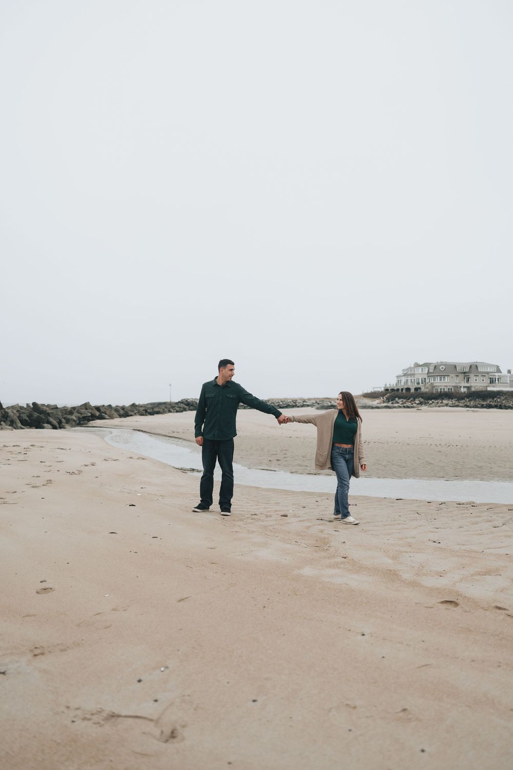 Couple holding hands and walking on a sandy beach on a cloudy day with a rocky jetty in the background.