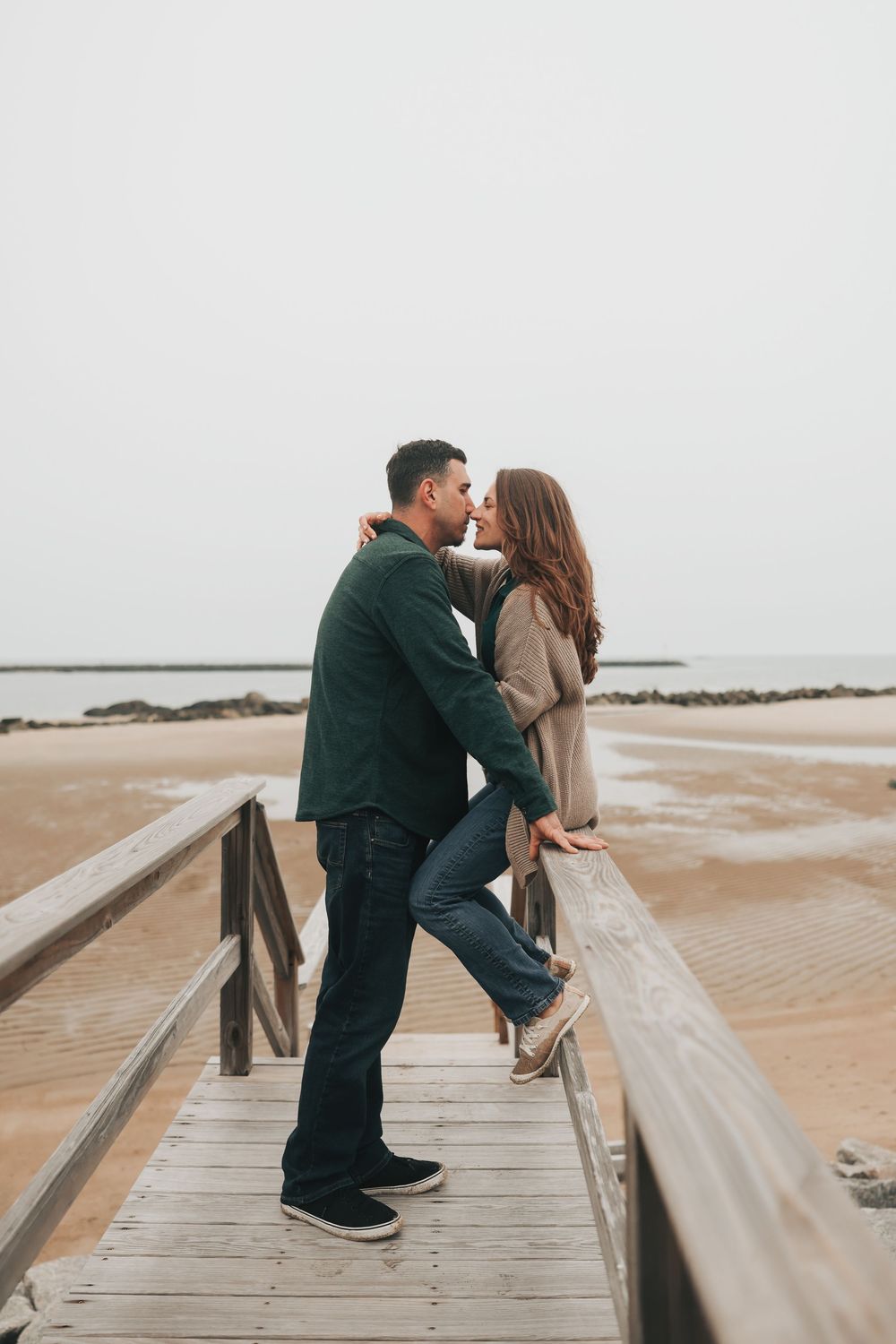Couple sharing a romantic kiss on a wooden boardwalk by the beach on a cloudy day.