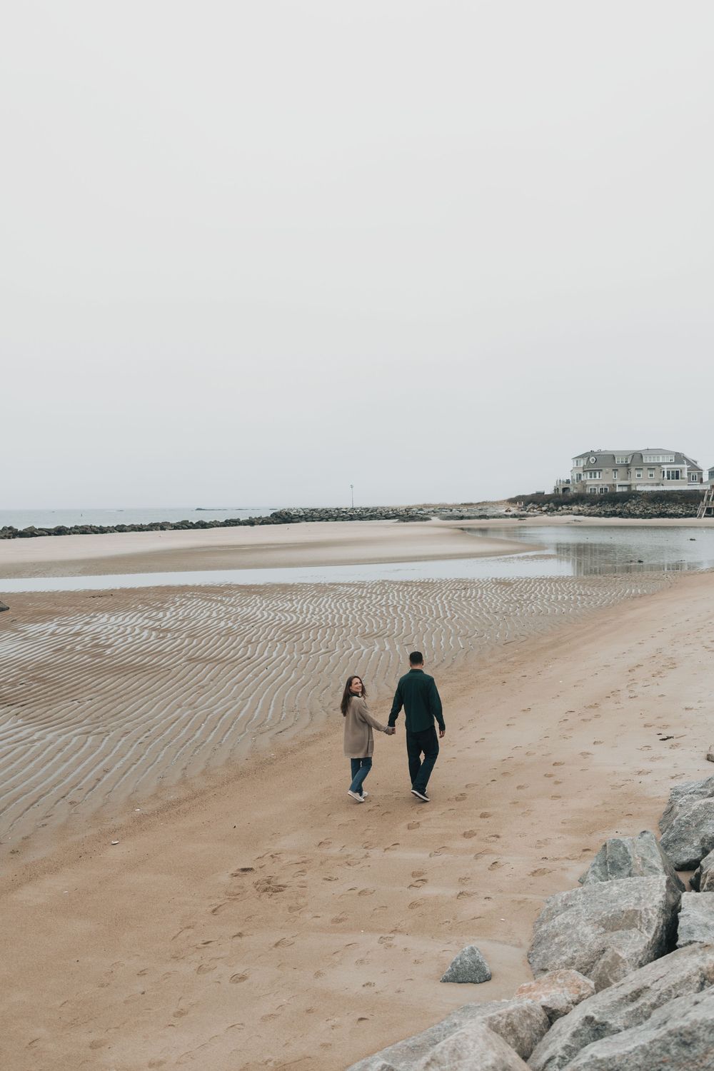 A couple holds hands while walking along a sandy beach on an overcast day, with rocky shores and a coastal house in the background.