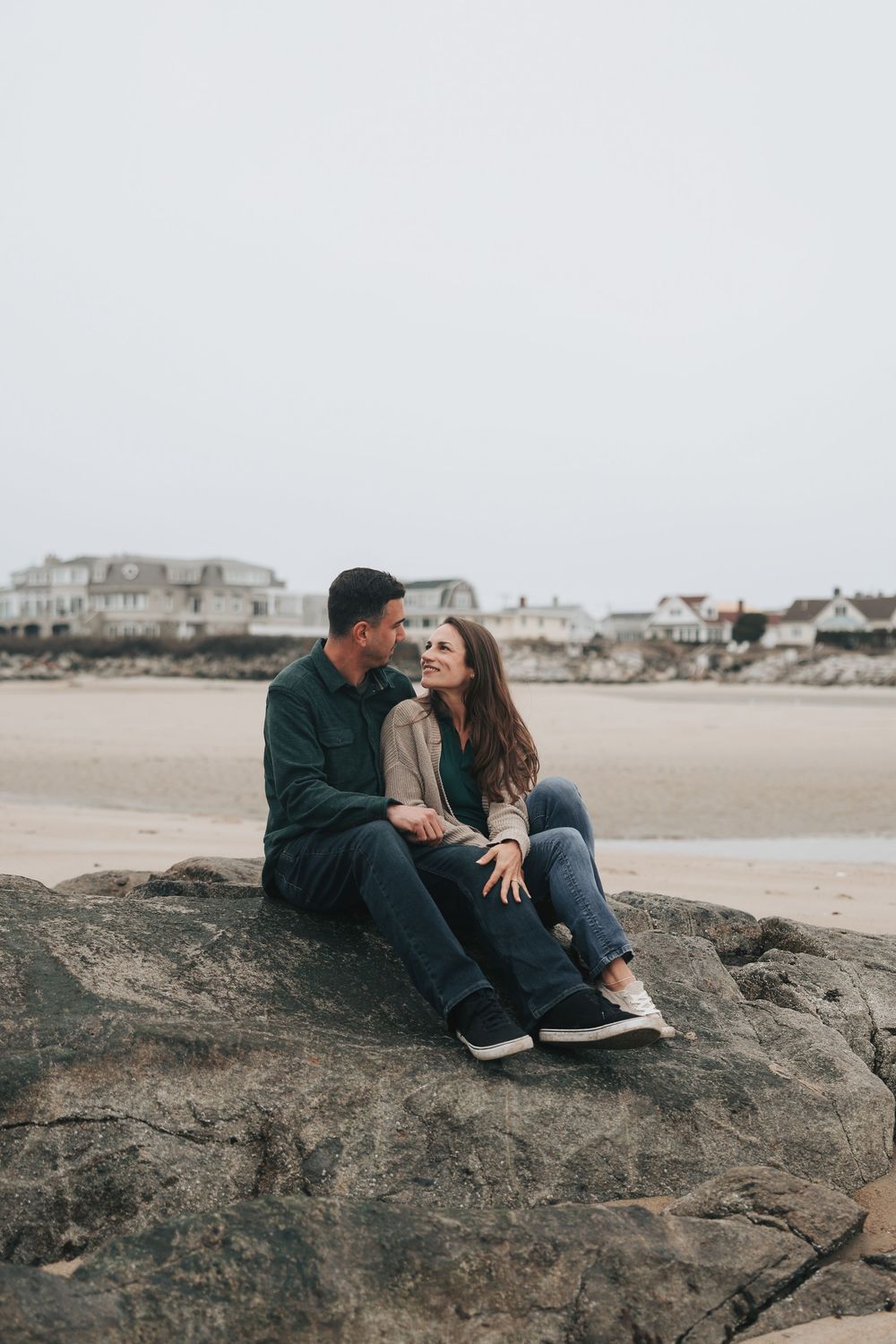 A couple sits on a rock at a sandy beach, smiling at each other on an overcast day.