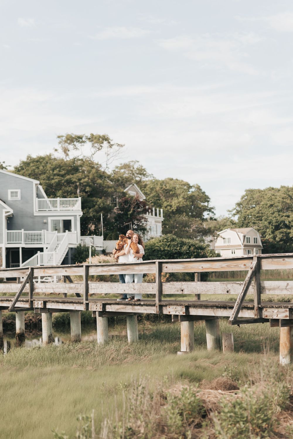 Couple standing on a wooden bridge with coastal New England homes and lush green trees in the background.