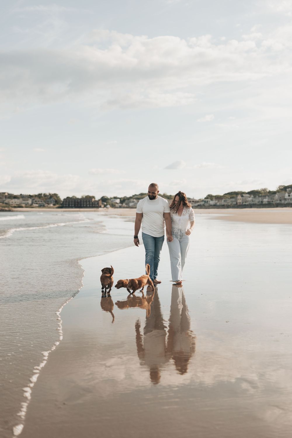 Couple walking two dogs on a wet reflective beach with calm waves and a cloudy sky backdrop.