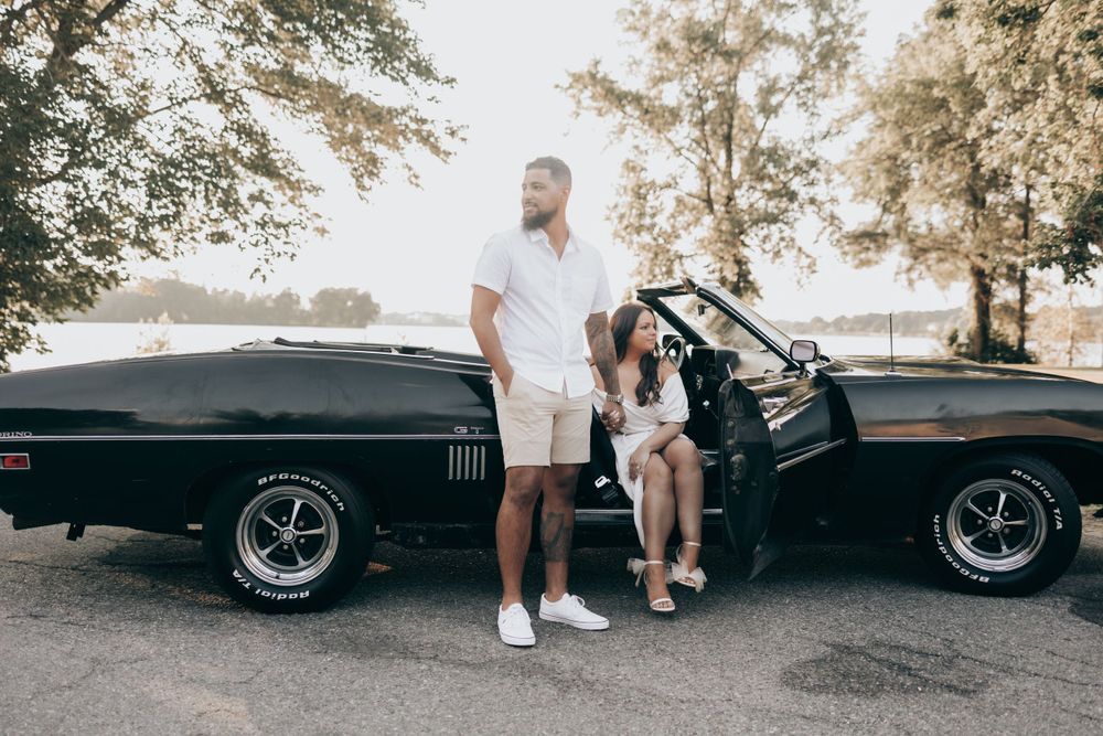 A couple poses with a black vintage muscle car under trees near water.