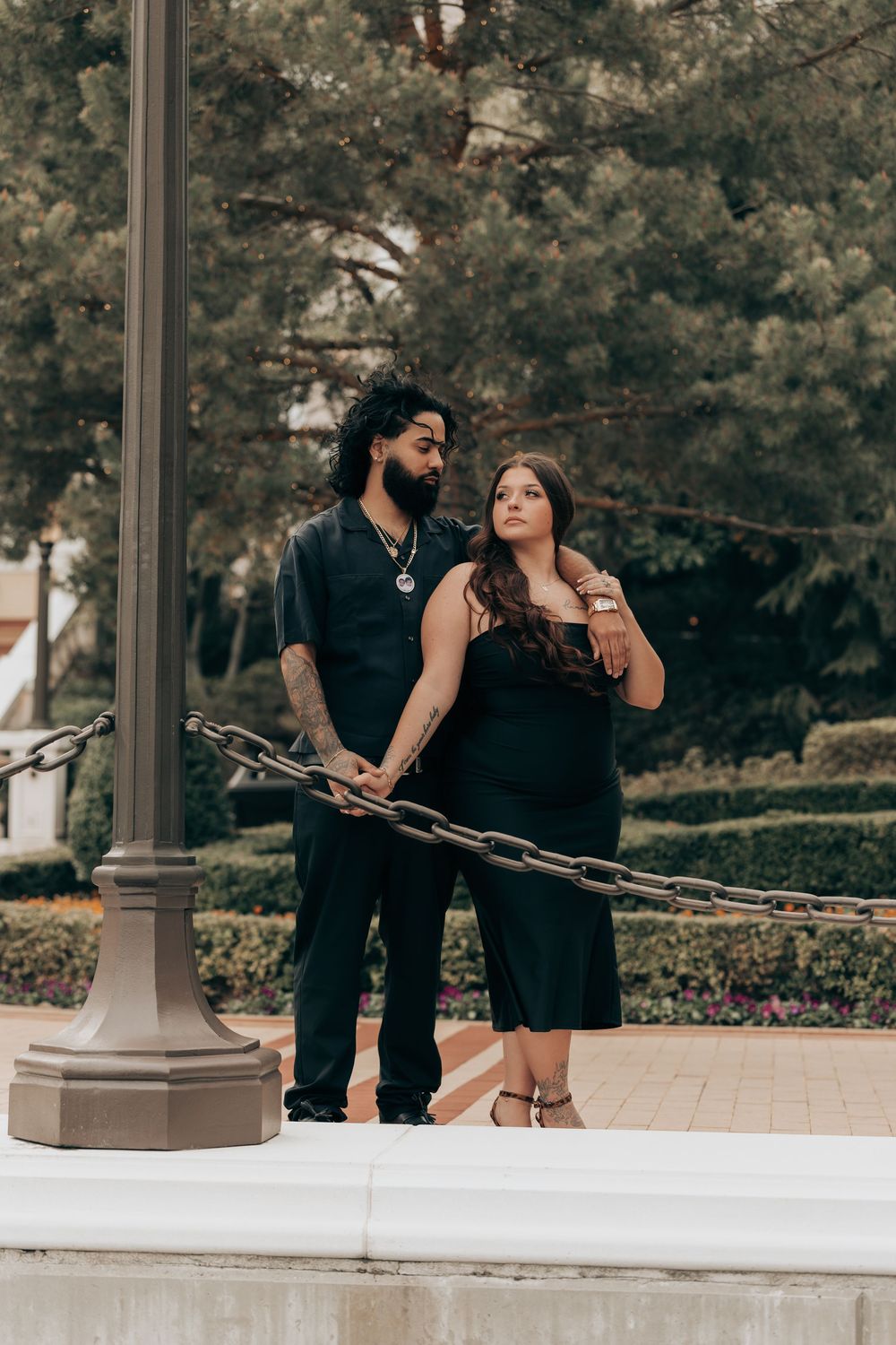 Couple in coordinating black outfits posing together near a lamp post in an outdoor garden setting at dusk.