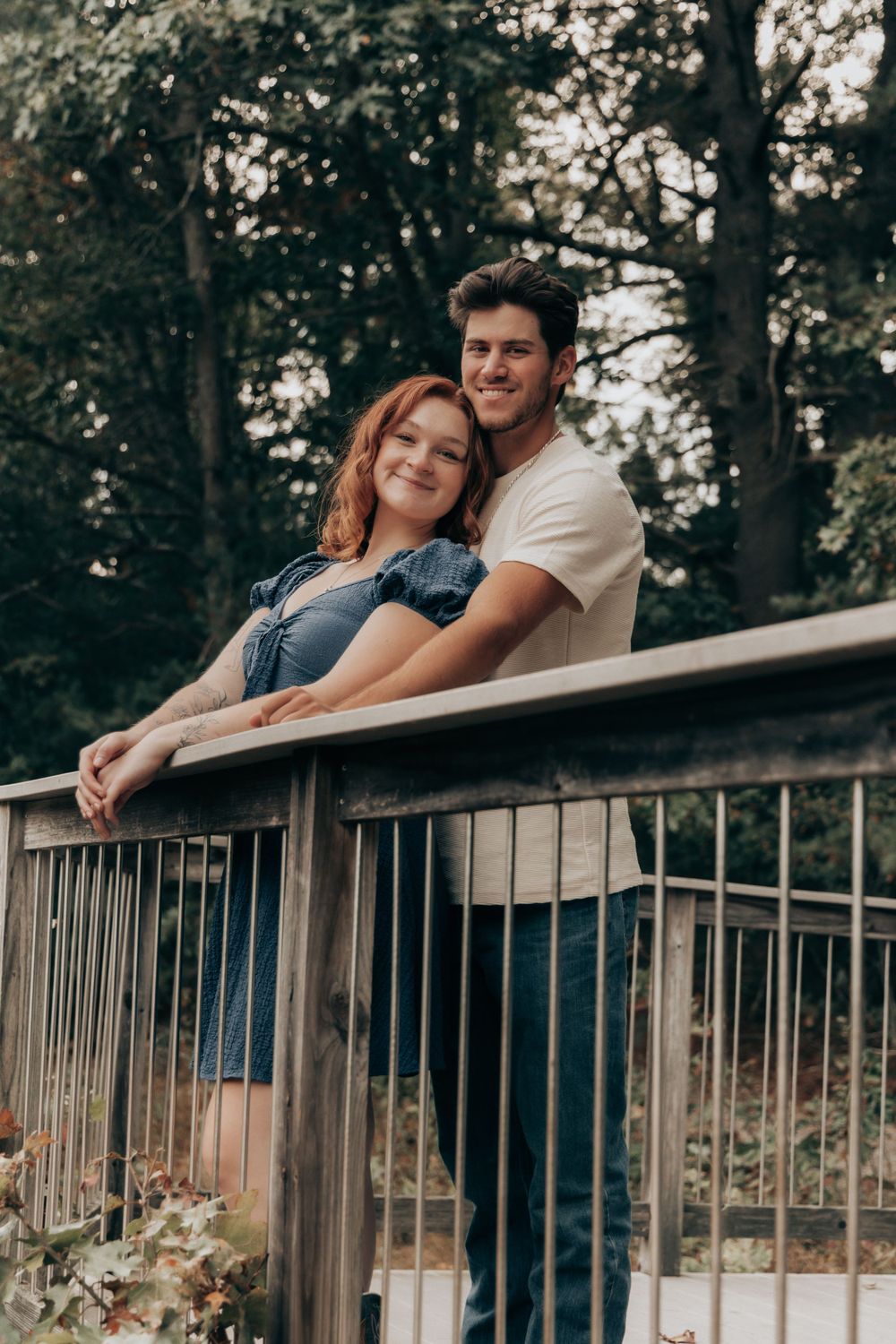 A couple leans against a wooden railing on a deck surrounded by trees.