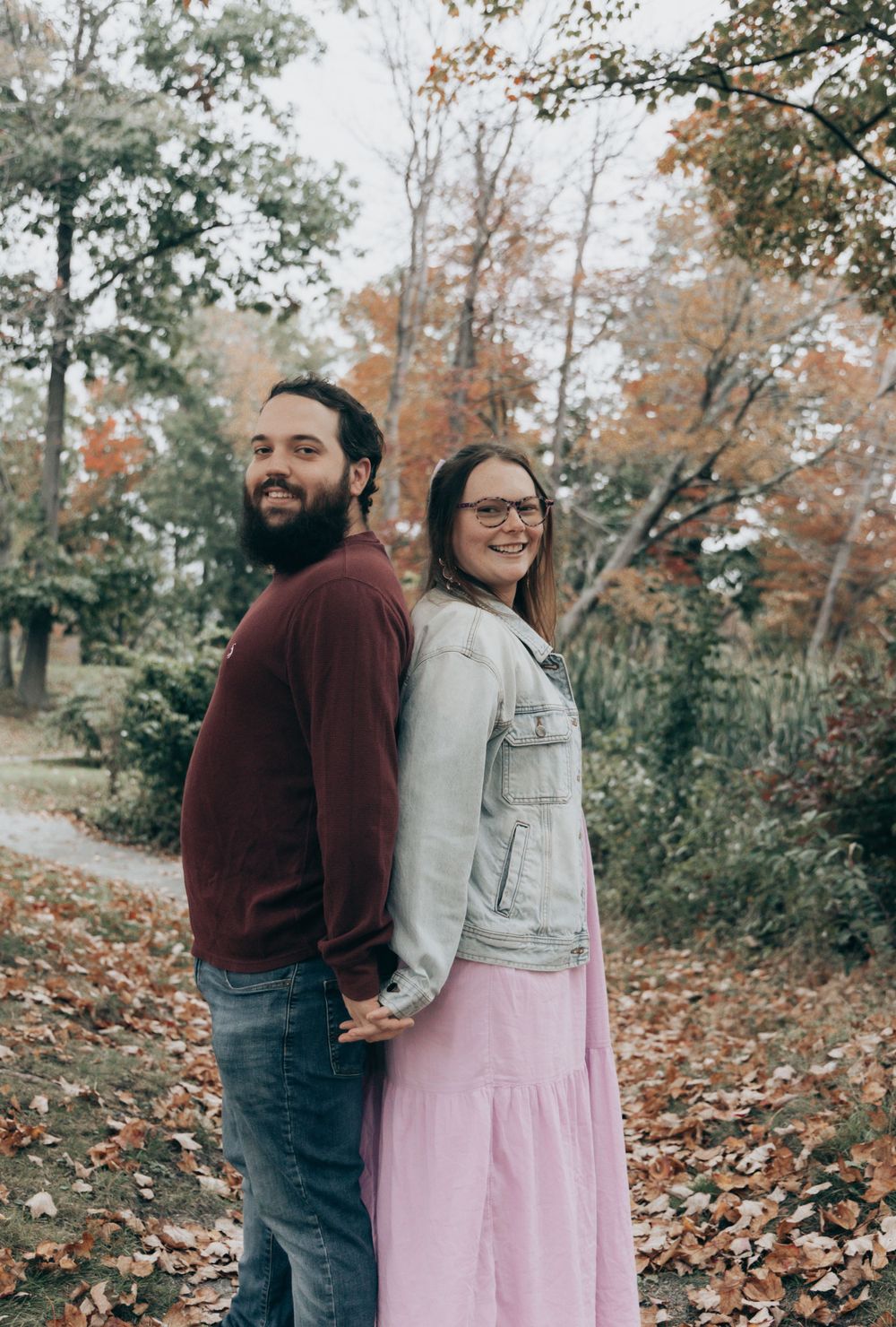 A couple stands back to back in an autumn forest setting wearing casual clothing against fall foliage.