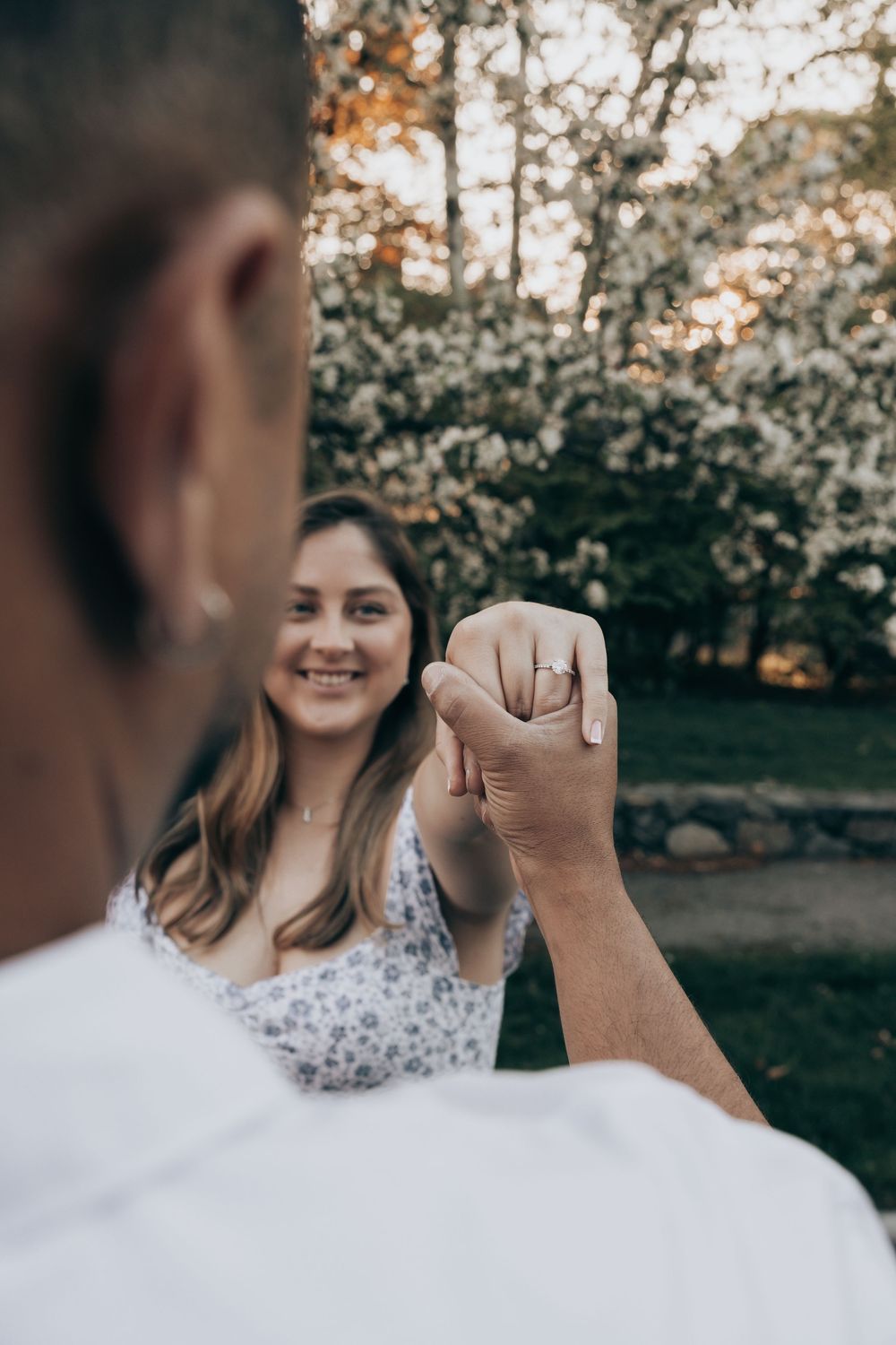 A couple holds hands in a romantic outdoor engagement setting with autumn trees in the background.