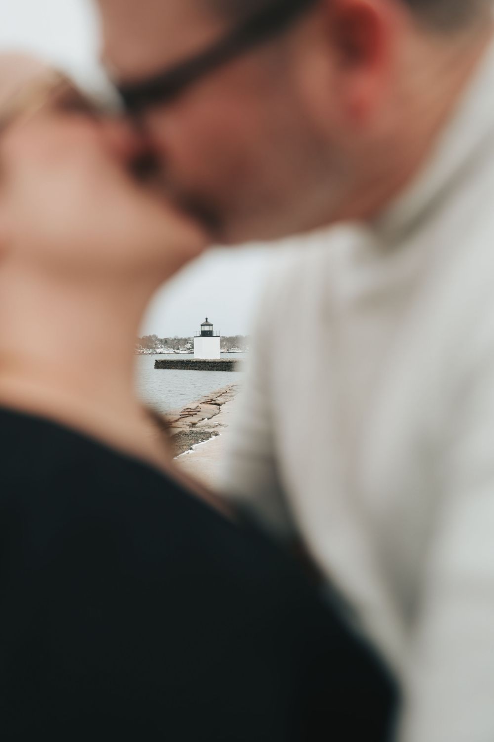 Couple kissing with a lighthouse visible between them in the blurred background near the water.