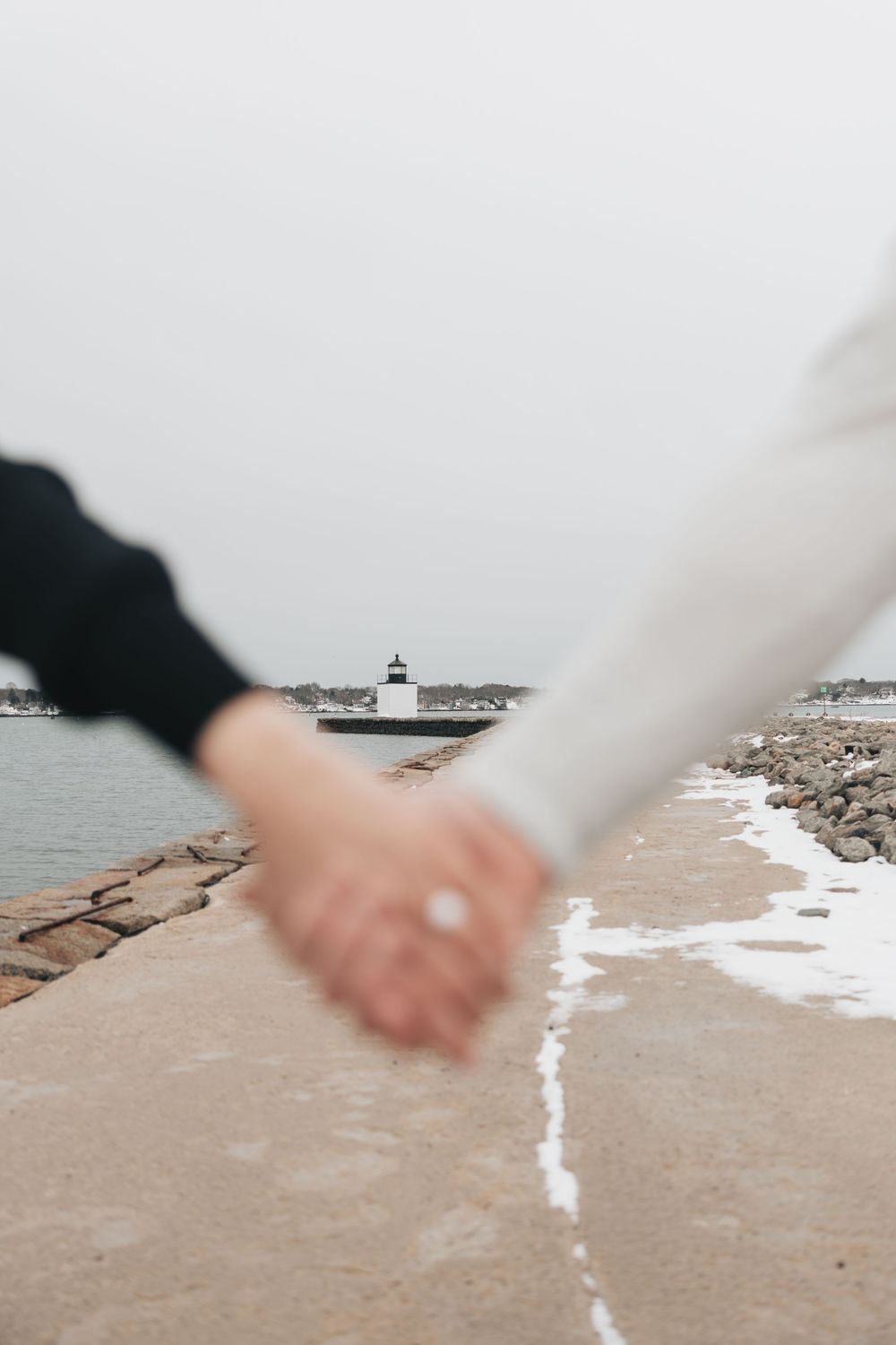 Couple holding hands on a snowy stone pier with a white lighthouse in the background on a cloudy day.