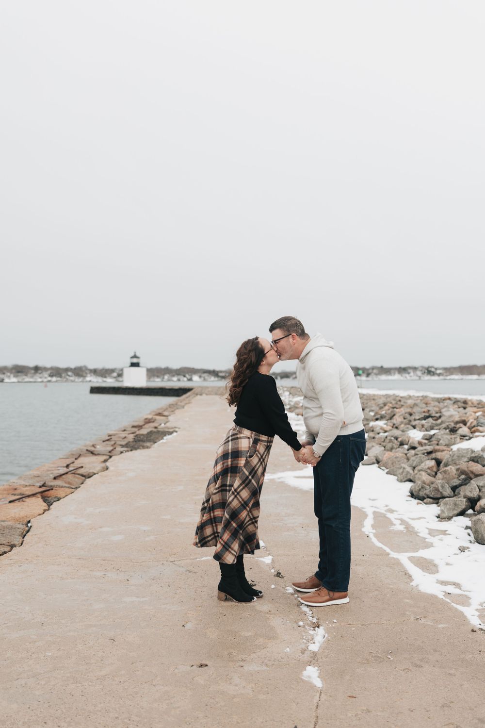 Couple sharing a romantic kiss on a snowy coastal pier with a lighthouse in the background on an overcast winter day.