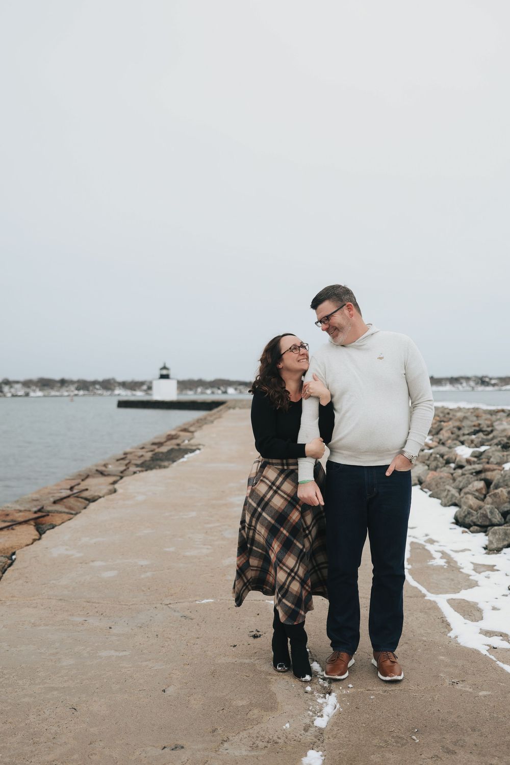Couple smiling at each other on a snowy coastal pier with a lighthouse in the background.