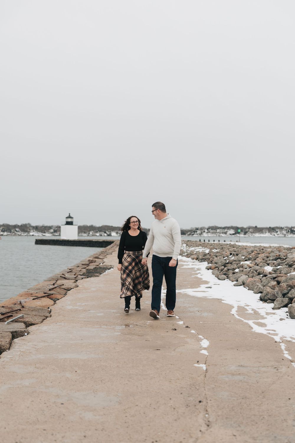 Couple walking hand-in-hand on a snowy coastal jetty with a lighthouse in the background on an overcast day.