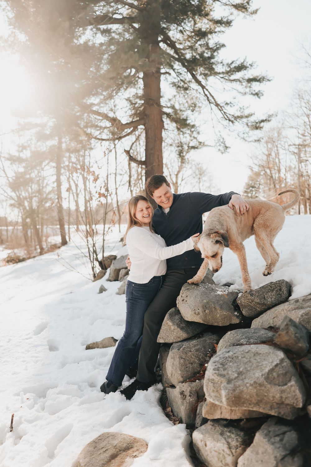 Couple smiling with golden dog on snowy rock wall surrounded by winter trees in bright sunlight.