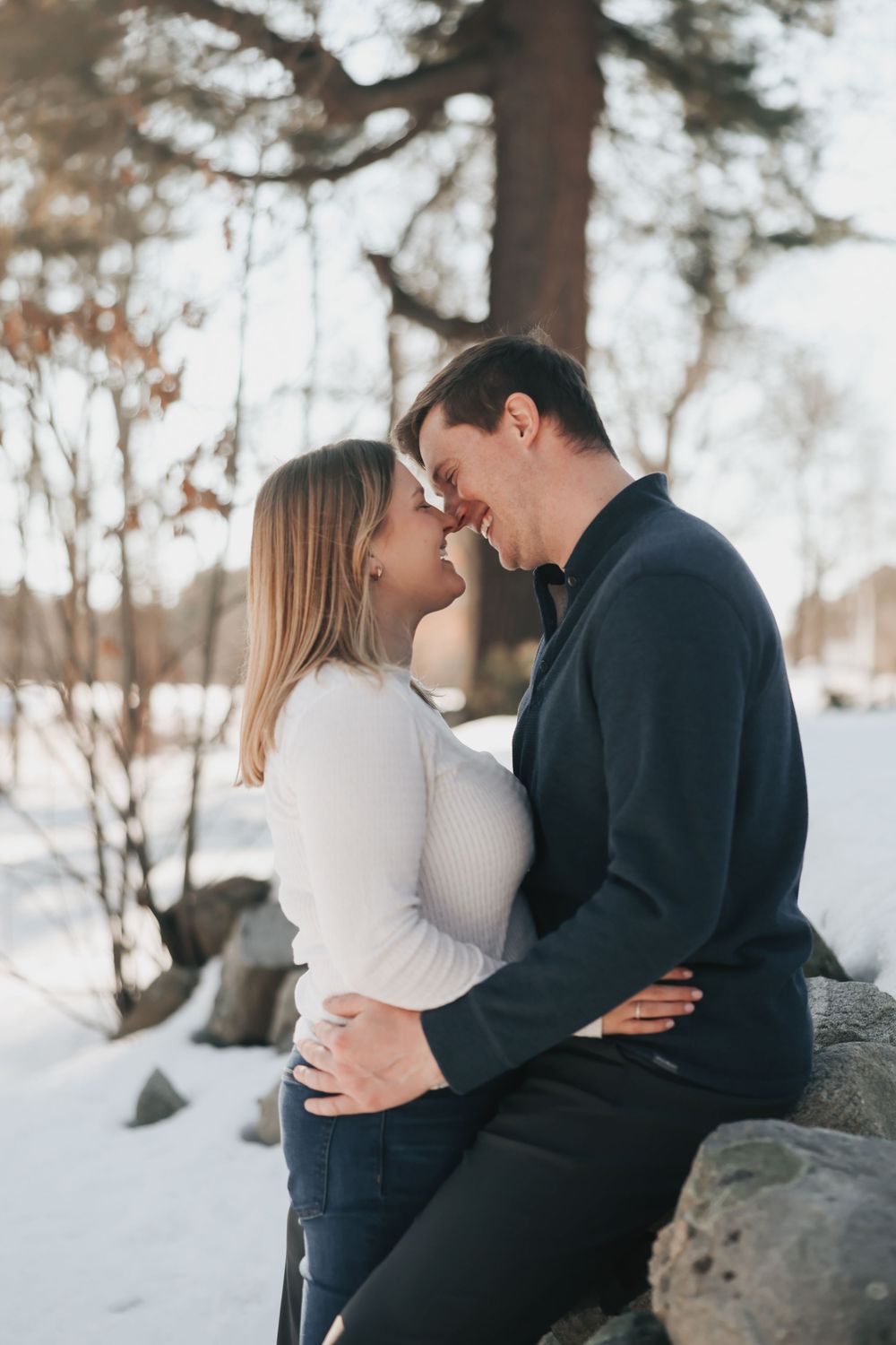 Couple sharing a romantic moment in a snowy winter forest, dressed in white and navy, sitting on rocks.