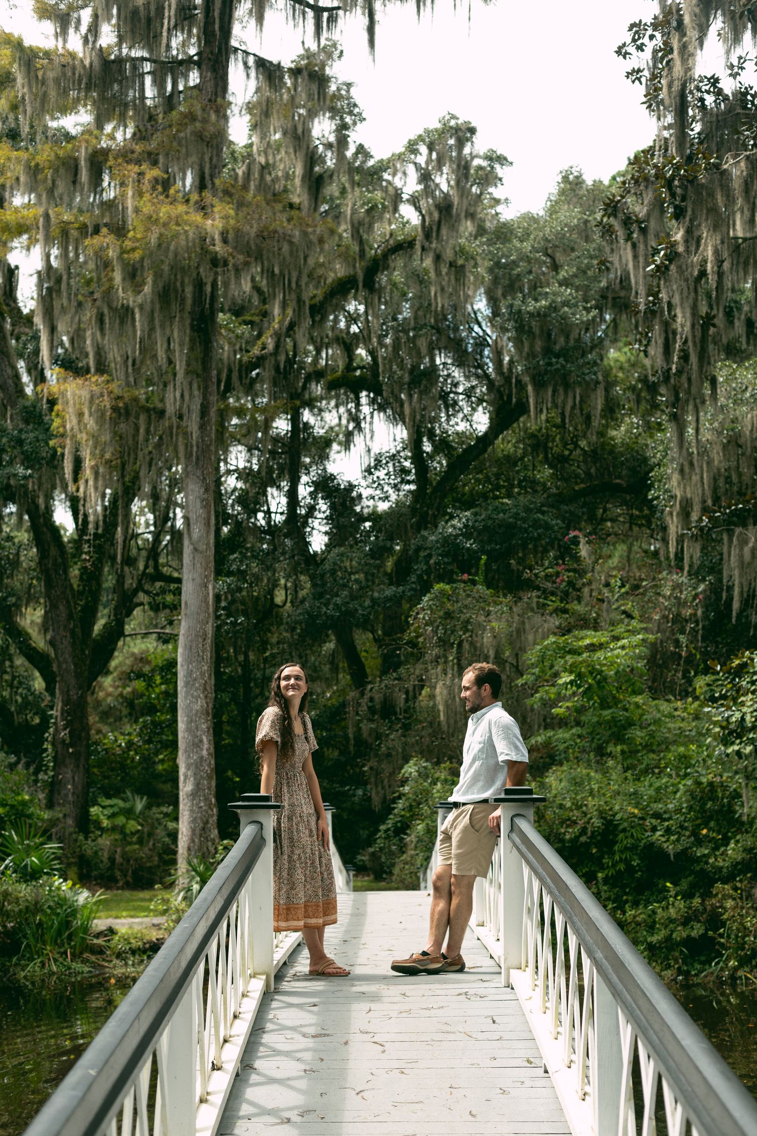 Two people stand together on a white bridge surrounded by Spanish moss draped trees.
