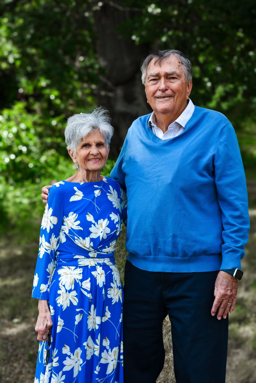 An elderly couple wearing coordinating blue outfits stand together outdoors against a green leafy background.