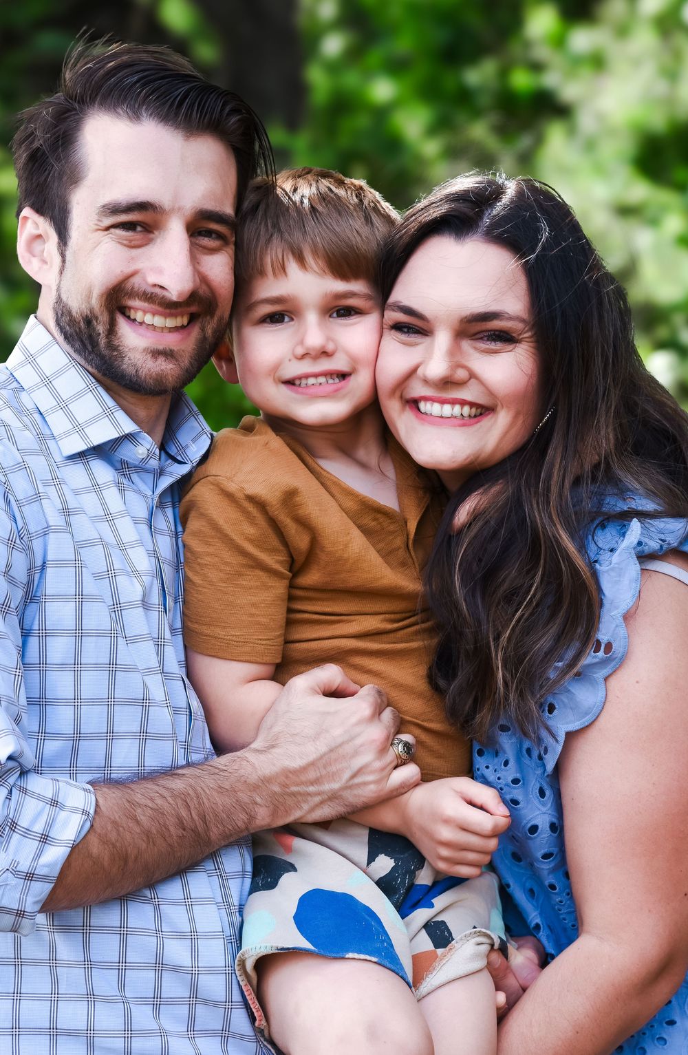 A happy family embraces and smiles together outdoors against a green leafy background.