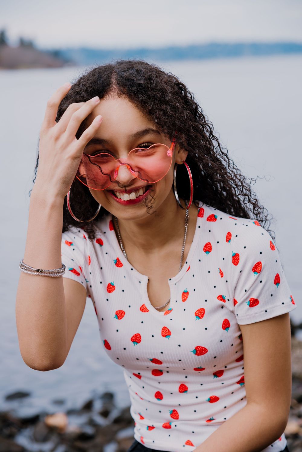 A person in a white and red polka dot top poses playfully near a waterfront.