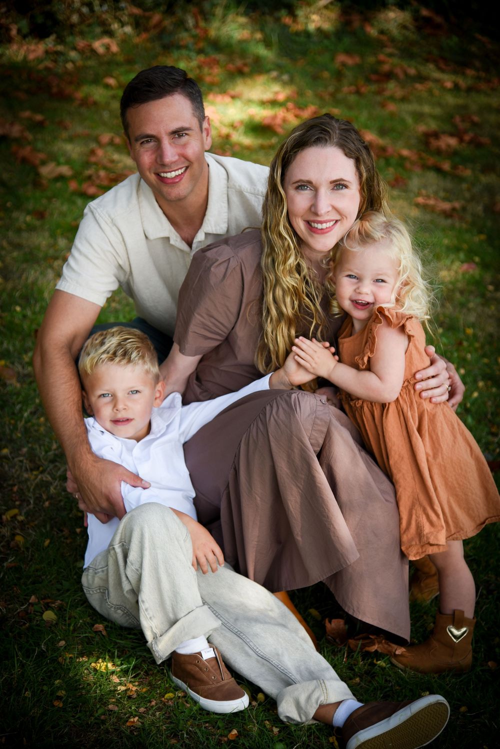 A smiling family of four poses together on a grassy lawn, with parents embracing a young boy and a little girl in an orange dress.