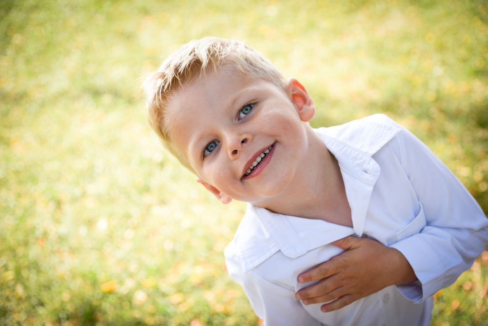 Smiling blond toddler boy in a white button-up shirt tilts his head while standing on sunlit green grass outdoors.