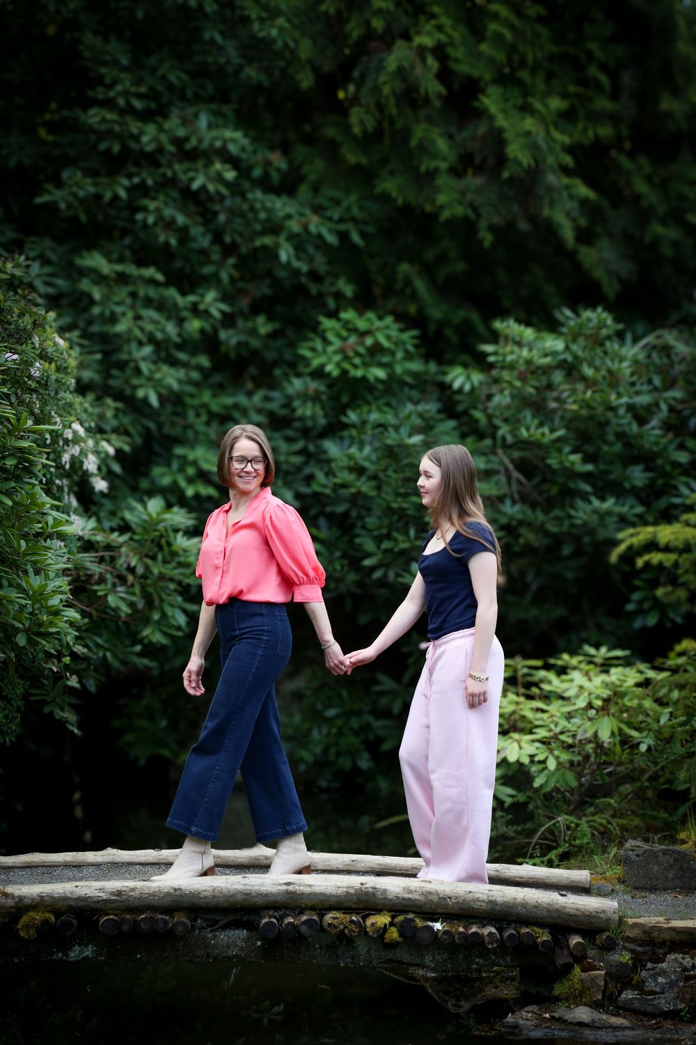 Two women holding hands on a wooden bridge surrounded by lush green trees in a garden setting.