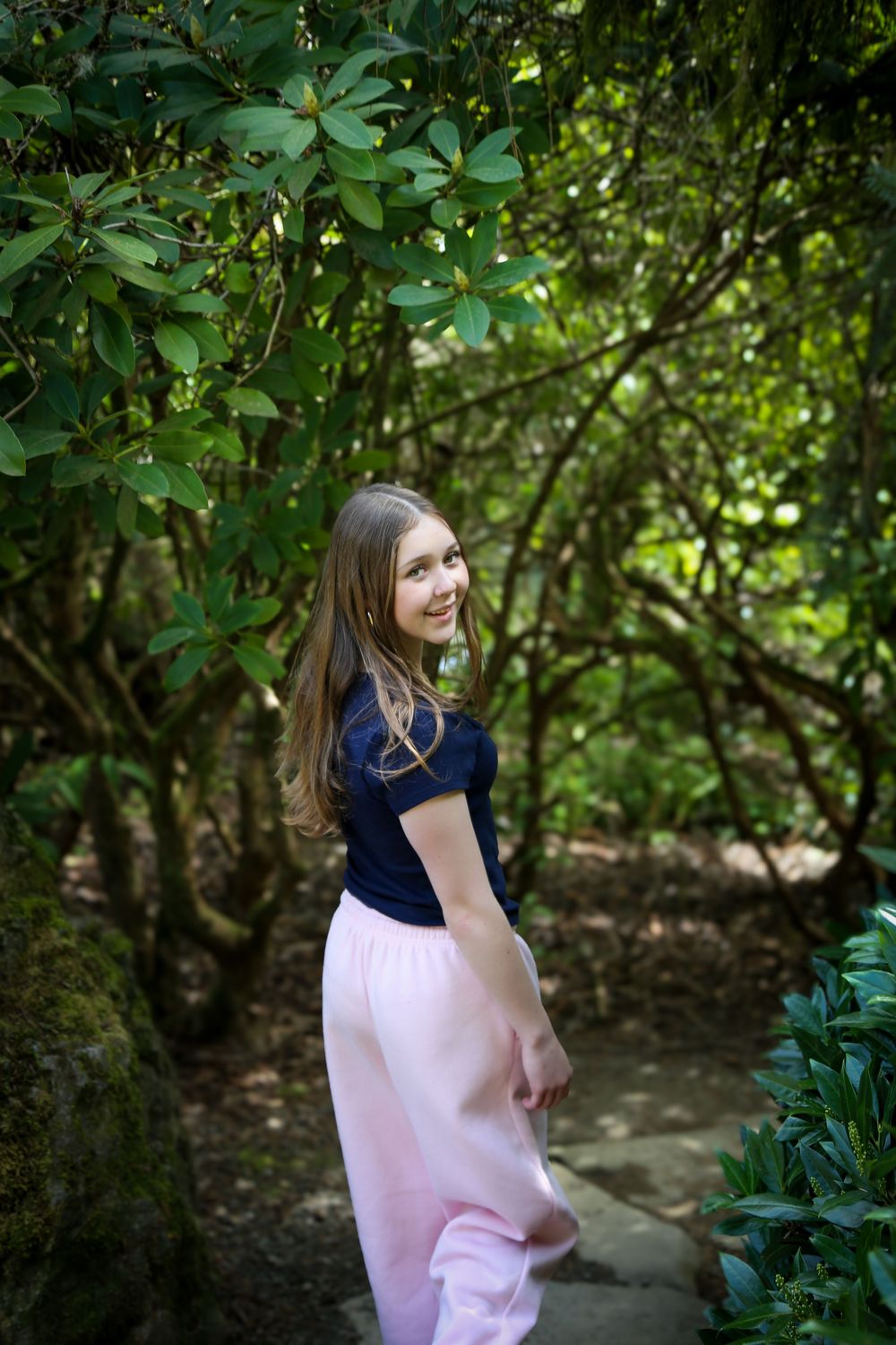 Young girl in pink skirt and navy top smiling in a lush green garden pathway surrounded by trees.