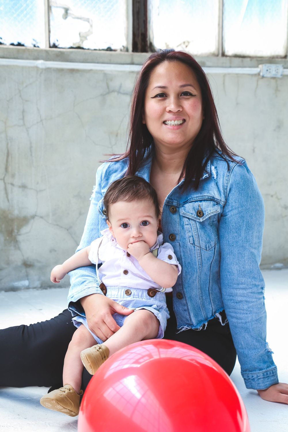 A smiling woman in a denim jacket sits on the floor holding a baby near a red balloon.