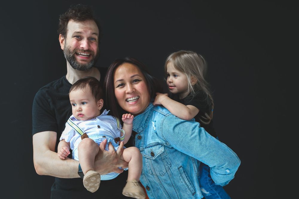Happy family portrait with parents holding baby and toddler girl, smiling against dark background.