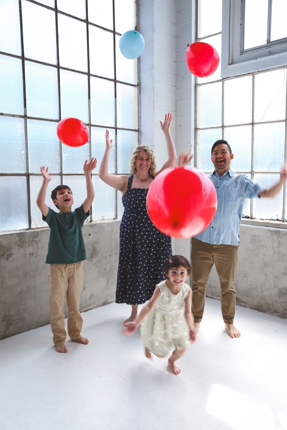 Family of four joyfully tossing red and blue balloons in a bright white studio with large windows.