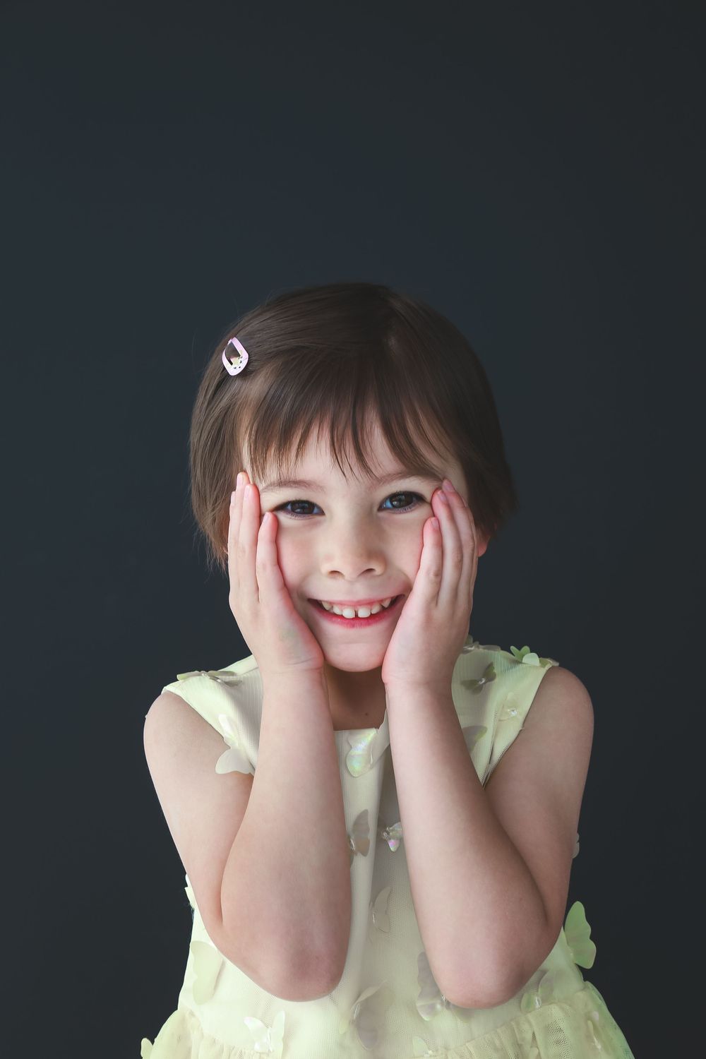 A joyful young girl in a yellow dress holds her cheeks with both hands, smiling brightly against a dark background.