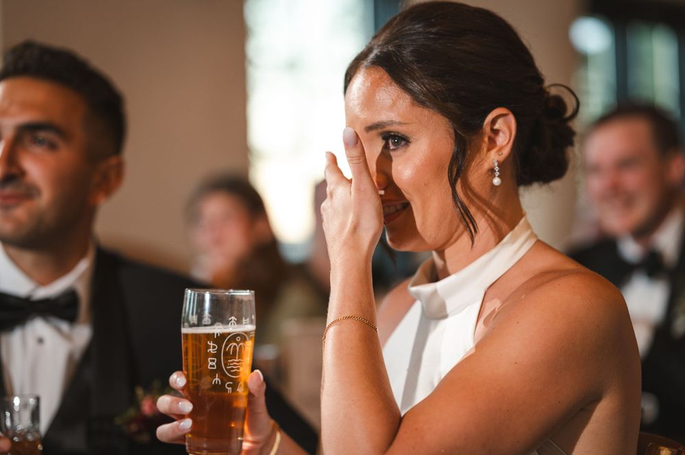 Bride holding a beer glass and wiping tears during speeches in industrial reception hall at Drowned Lands Brewery in Warwick, NY.