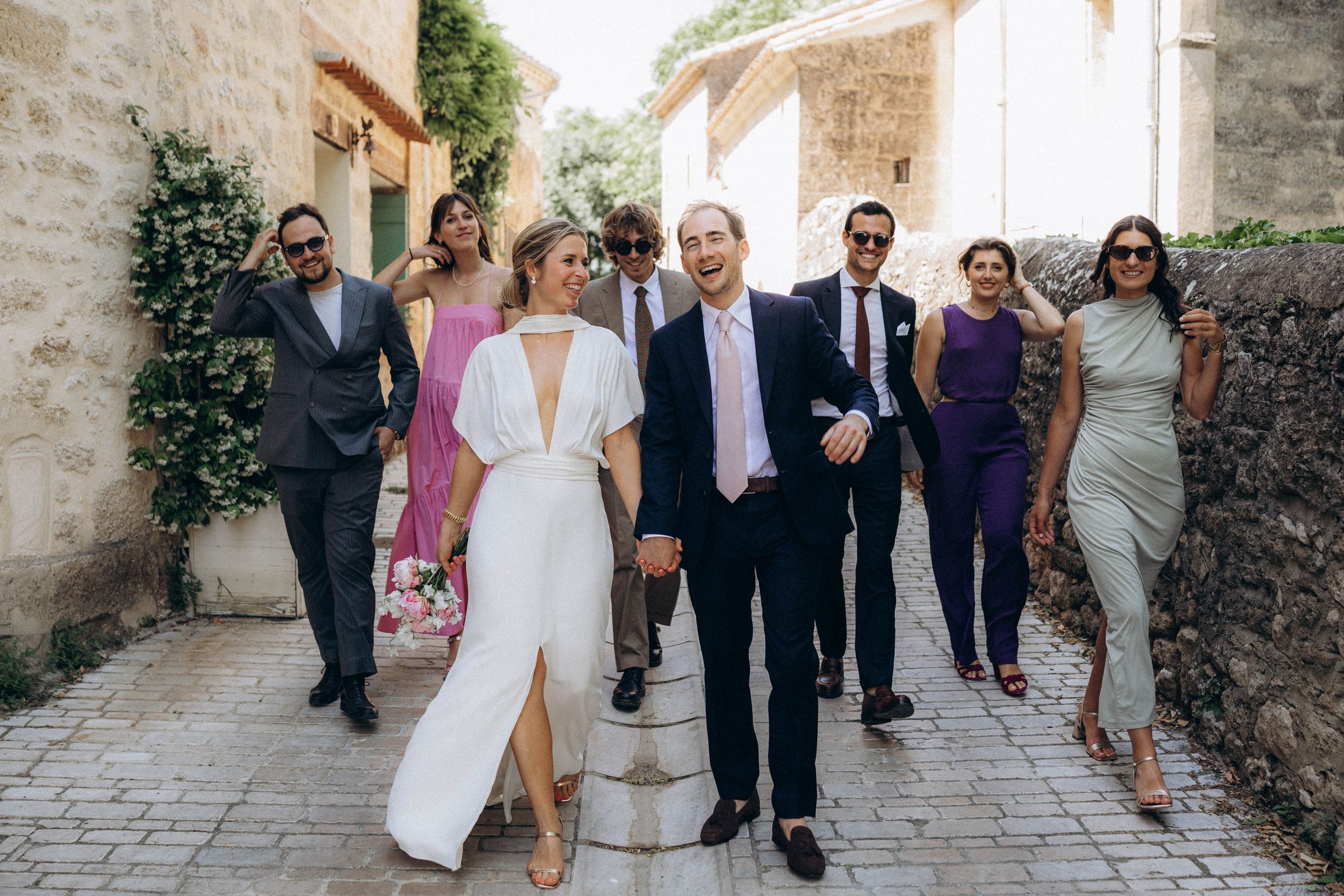 Wedding party walks cobblestone alley, bride in white gown, groom in suit, guests in colorful formal attire.