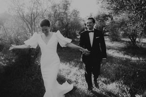 Black and white photo of a bride and groom walking hand in hand through an olive grove on their wedding day.