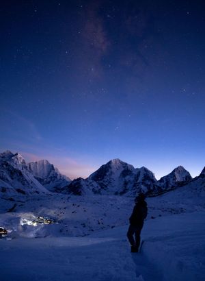 Starlit winter night scene of snow-covered mountains with distant lights in the valley.