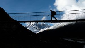 Suspension bridge spans across a mountain valley with snow-capped peaks in the background.