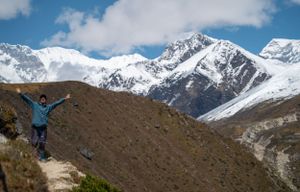 Hikers traverse a mountain trail with snow-capped Himalayan peaks in the background.